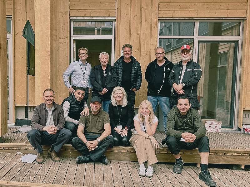 En gruppe kolleger og håndværkere sidder og står på terrassen ved et nyt træhus.