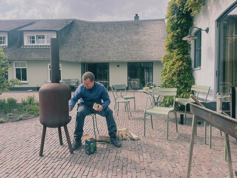 En kollega tænder en udendørs brændeovn i gården ved et stråtækt hus.